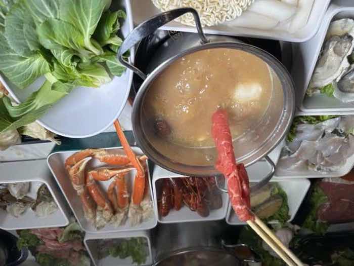 Chopsticks lowering a rolled slice of beef into a simmering golden broth beside plates of crab legs and crawfish