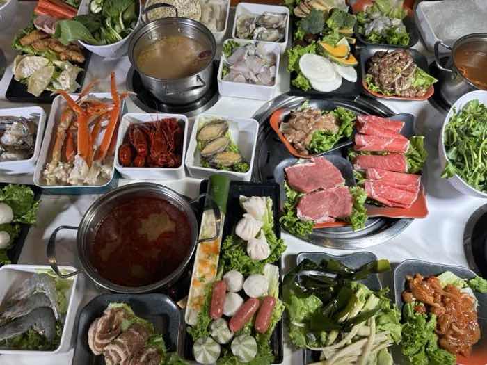 Overhead view of a full table with red and clear broths, snow crab legs, sliced beef, pork belly and seafood around a central grill