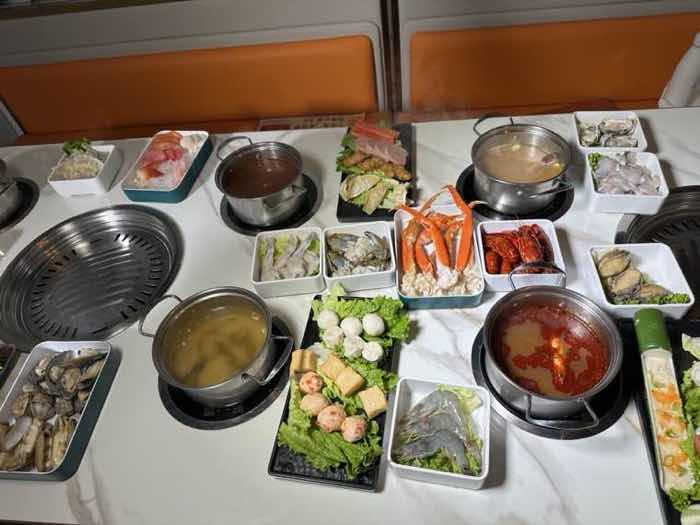Overhead view of a booth table with a built-in grill, three broth pots, crab legs, clams, sliced fish and vegetables