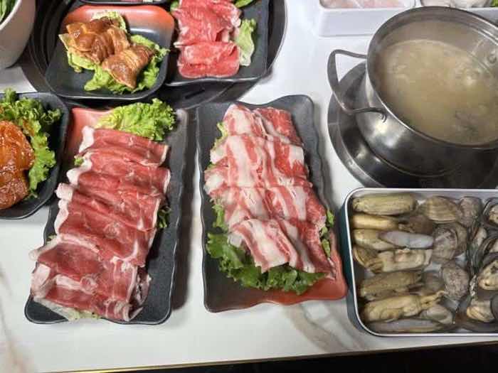 Platters of thinly sliced beef and pork belly beside a tray of fresh clams and a pot of clear broth