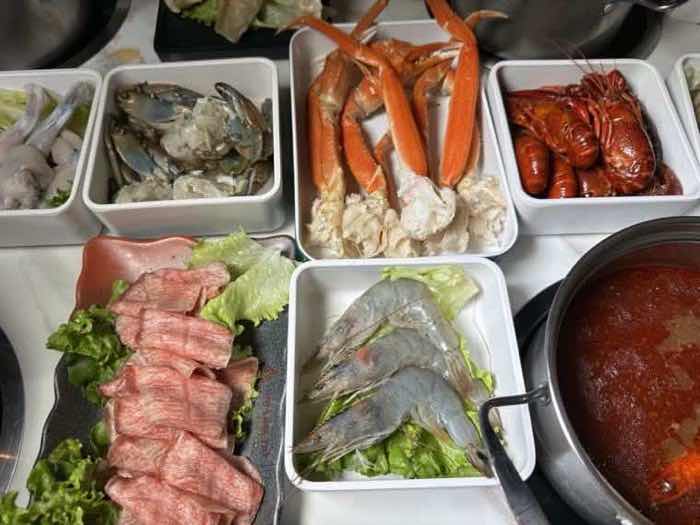 Snow crab legs, crawfish, blue crab and raw prawns plated beside marbled beef and a pot of spicy red broth